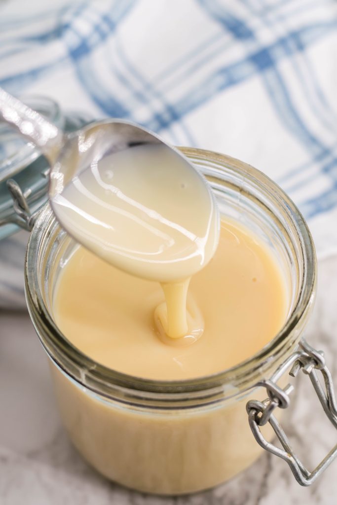a silver spoon shown with homemade sweetened condensed milk dripping from it into a glass jar