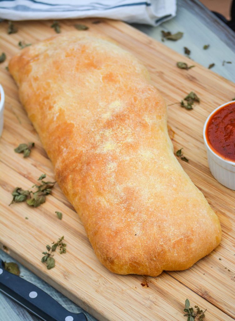 a crispy golden brown crusted meat & cheese stromboli shown on a wooden cutting board surrounded by herbs with a bowl of marinara sauce off to the side