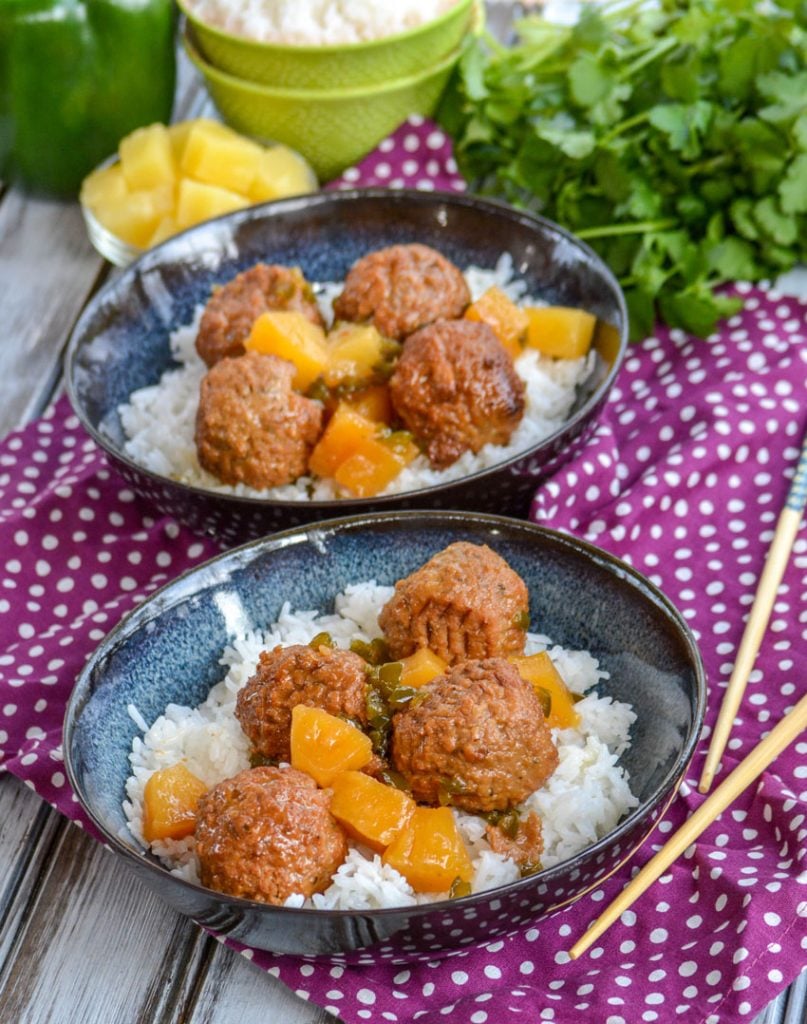 Crockpot Hawaiian Meatballs over a bed of rice in glazed bowls on a purple napkin with chop sticks and fresh herbs in the back ground