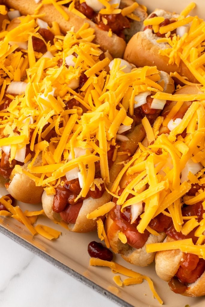 hot dogs in a casserole dish topped with shredded cheddar cheese and diced white onions