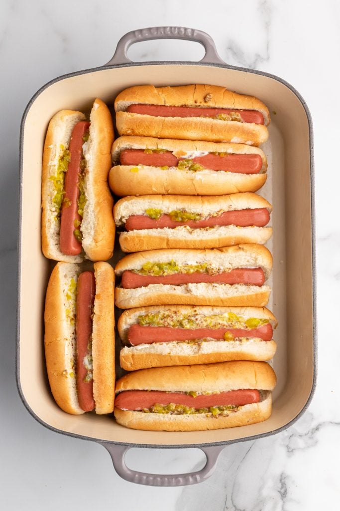 raw hot dogs in buns spread with condiments arranged in a casserole dish