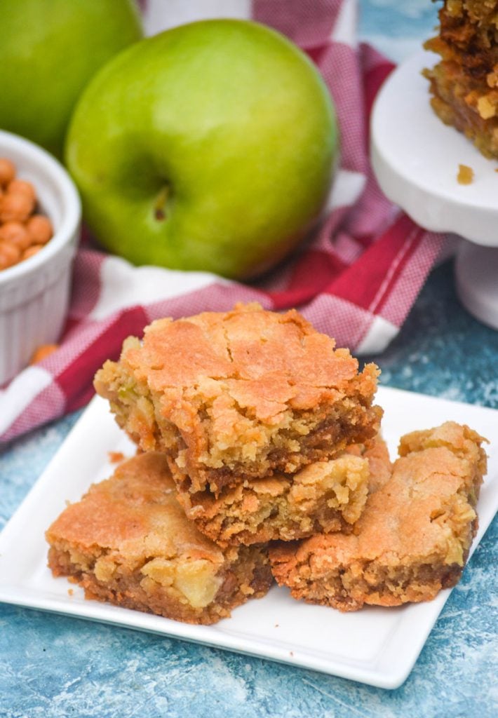 a stack of caramel apple blondies on a white square appetizer plate with whole apples in the background