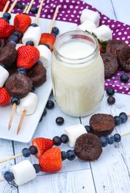 dessert kabobs on wooden skewers shown on a white platter and wooden background with a glass of milk and fresh berries in the background