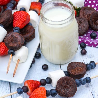 dessert kabobs on wooden skewers shown on a white platter and wooden background with a glass of milk and fresh berries in the background