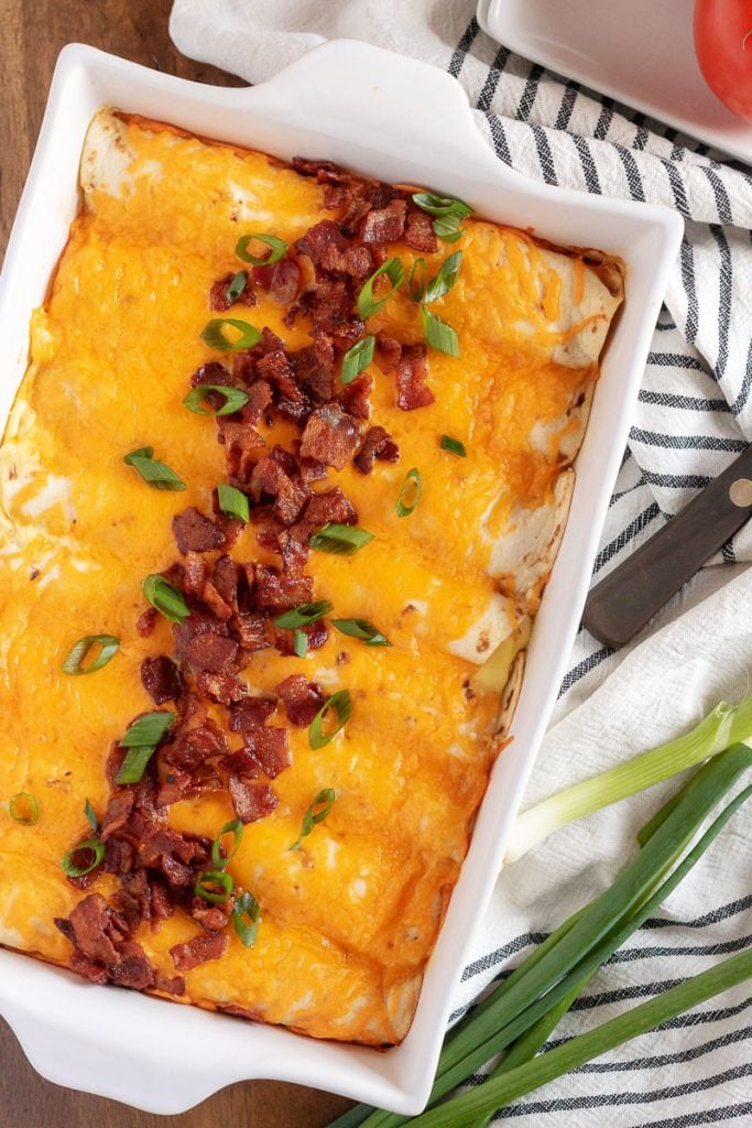 a white casserole dish filled with cheesy breakfast enchiladas garished with crisp crumbled bacon and green onions