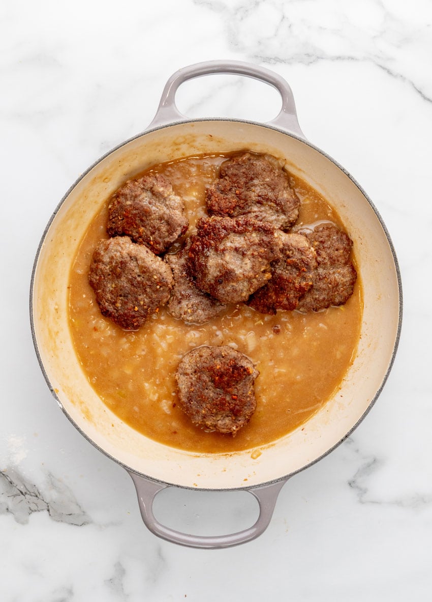 homemade hamburger steak patties frying in onion gravy in a large enameled cast iron skillet