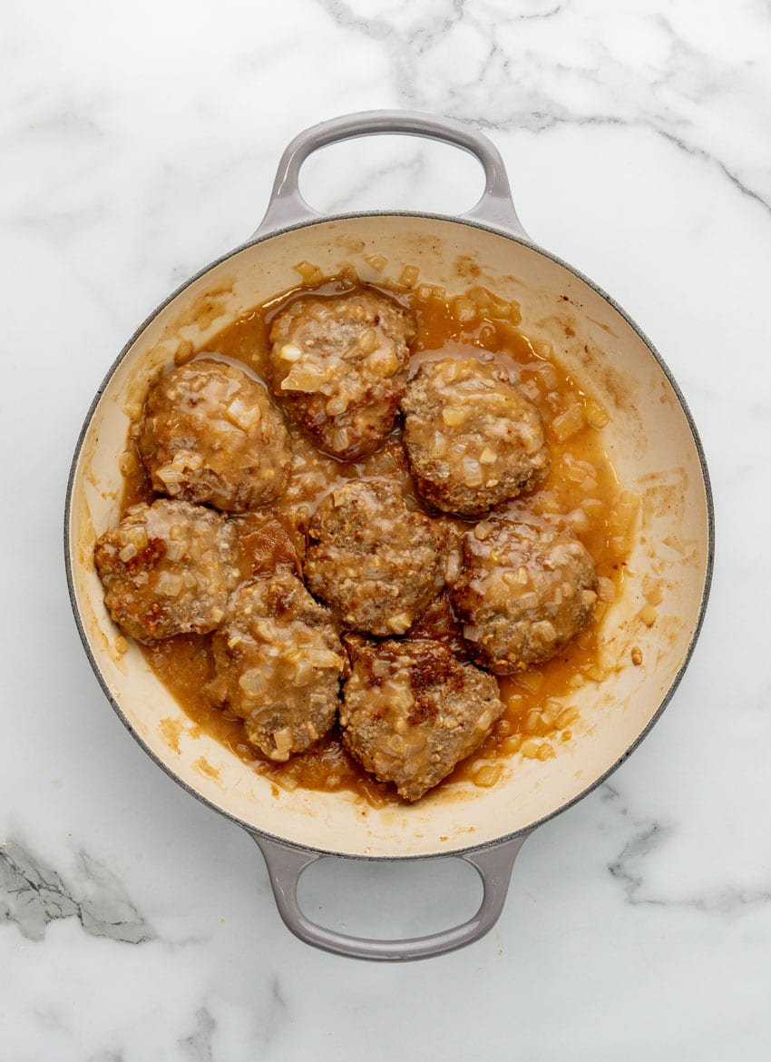 homemade hamburger steak patties frying in onion gravy in a large enameled cast iron skillet