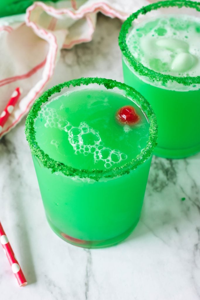 Christmas grinch punch served in green sugar rimmed glasses with a maraschino cherry floating in the green liquid