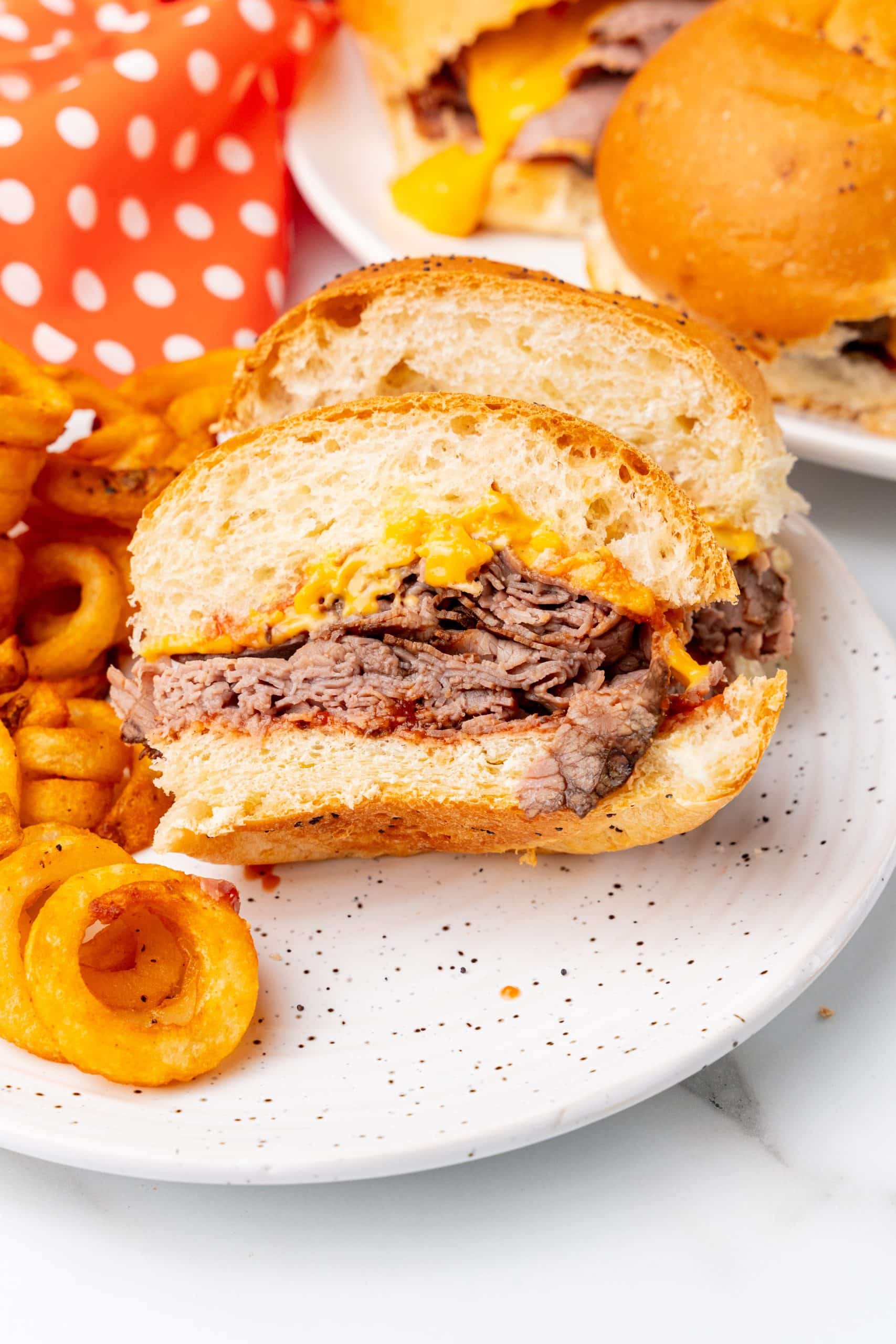 two halves of a homemade arby's roast beef sandwich with cheese sauce on a speckled white plate with curly fries on the side