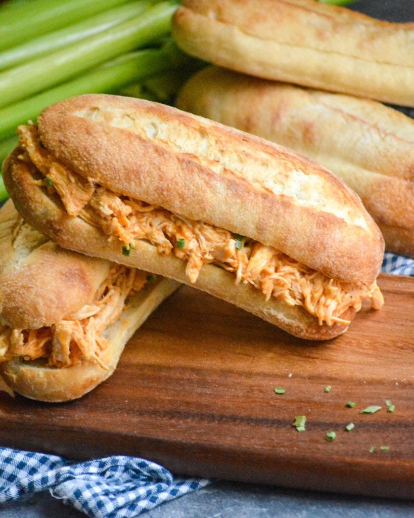 crockpot buffalo chicken sandwiches shown stacked on a wooden cutting board with rolls and crisp celery stalks in the background