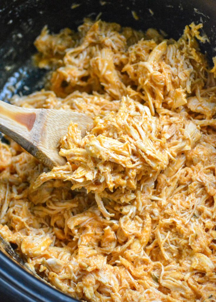 a wooden spoon shown stirring crockpot buffalo chicken in the black bowl of a pot