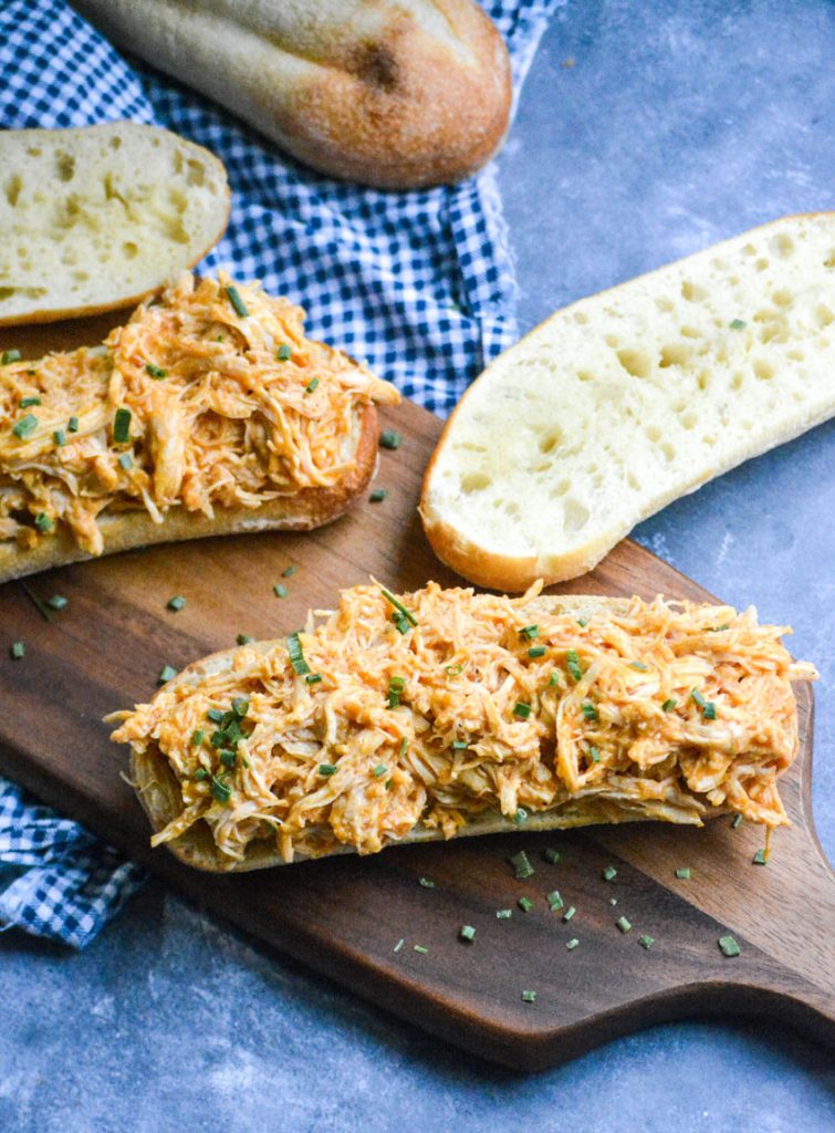 crockpot buffalo chicken sandwiches shown open faced on a wooden cutting board on top of a white & blue checkered napkin