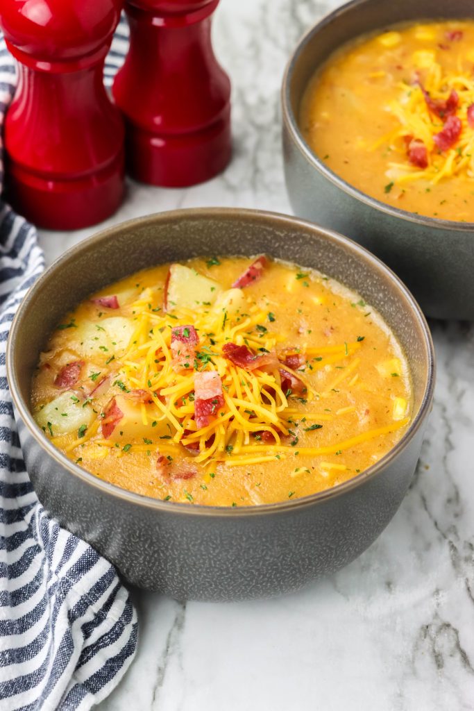 corn and potato studded smoky bacon and cheddar chowder shown in gray soup bowls topped with shredded cheddar cheese and crisp bacon bits