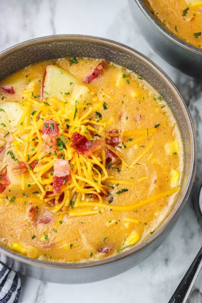 corn and potato studded smoky bacon and cheddar chowder shown in gray soup bowls topped with shredded cheddar cheese and crisp bacon bits