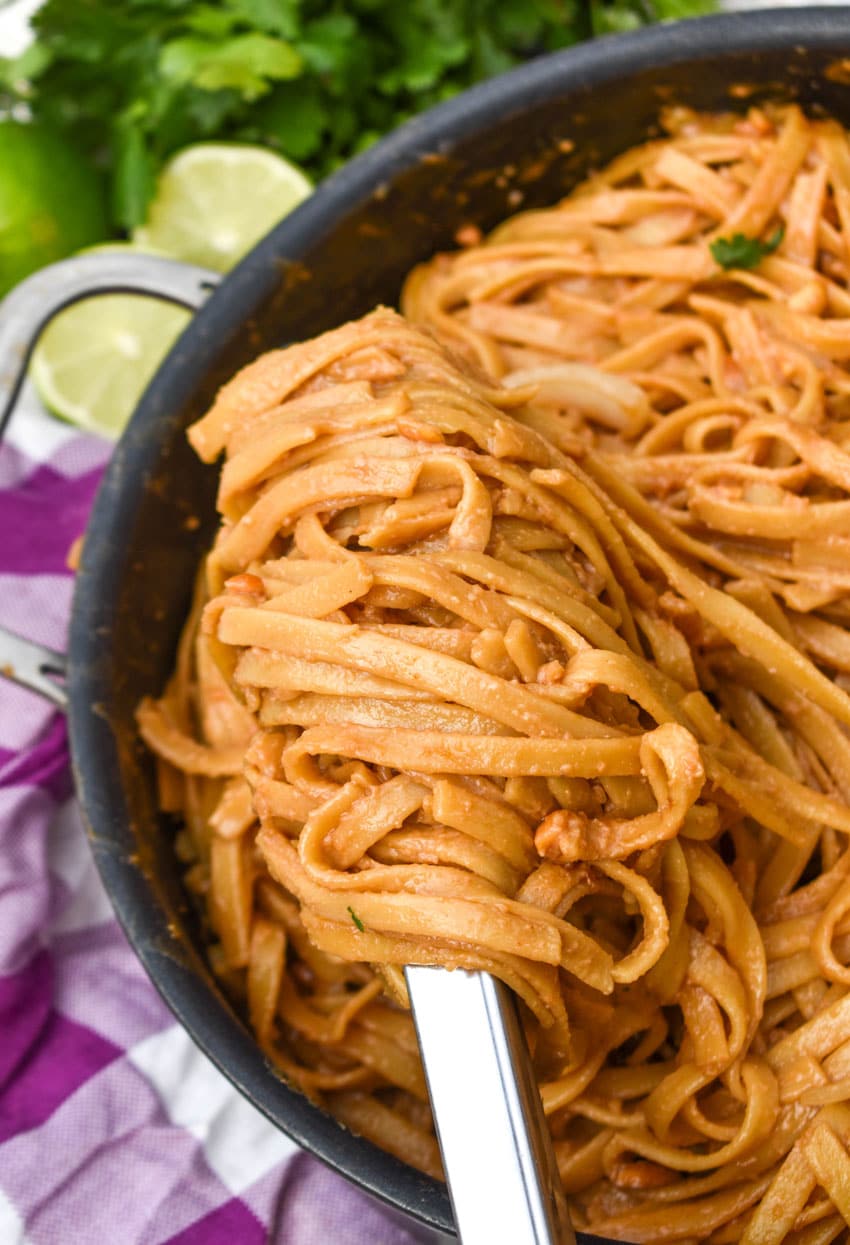 a silver ladle scooping peanut butter noodles out of a large black skillet