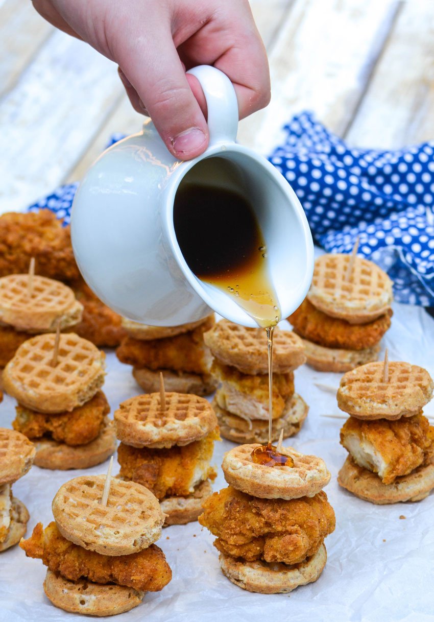maple syrup being poured from a white pitcher over chicken and waffle sliders