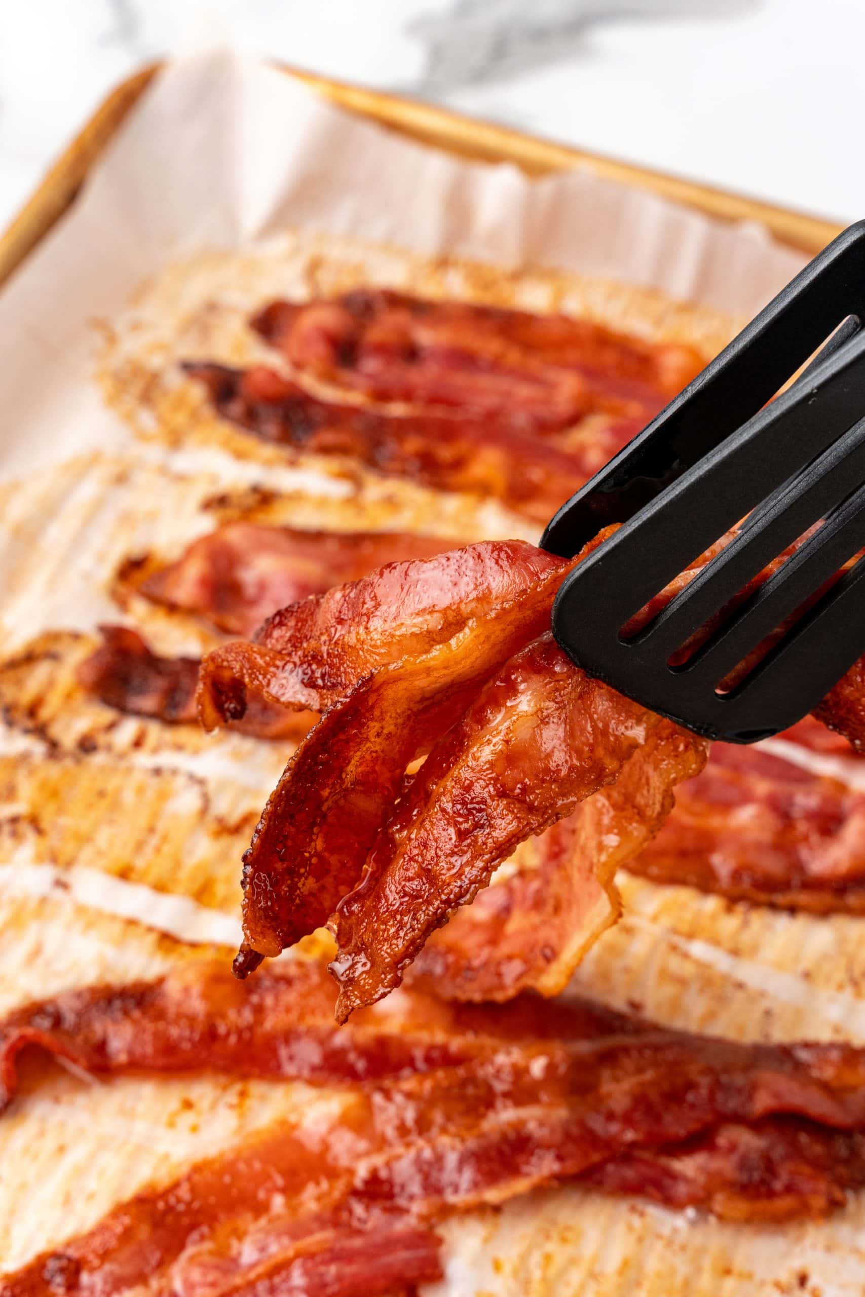 tongs holding up crispy slices of baked bacon in the oven