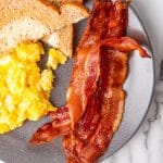crispy strips of oven baked bacon on a gray plate next to scrambled eggs and toast