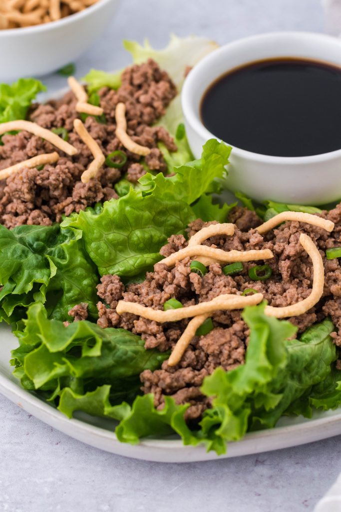 sriracha beef lettuce wraps topped with crispy chow mein noodles and served on a white plate with a cup of soy sauce for dipping