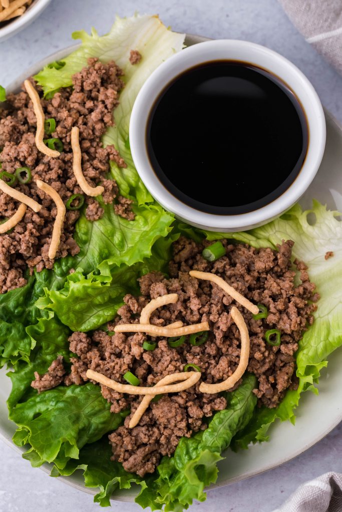 sriracha beef lettuce wraps topped with crispy chow mein noodles and served on a white plate with a cup of soy sauce for dipping