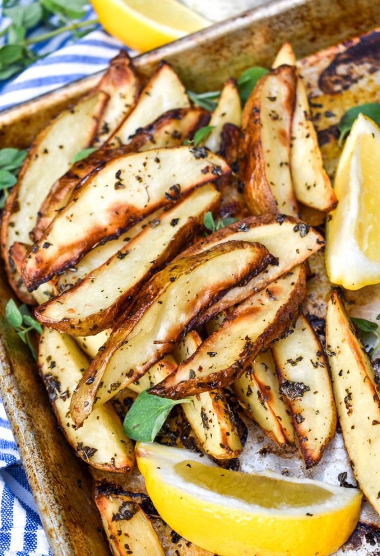 a pile of Greek oven fries in the corner of a baking sheet next with fresh lemon wedges and oregano l eaves