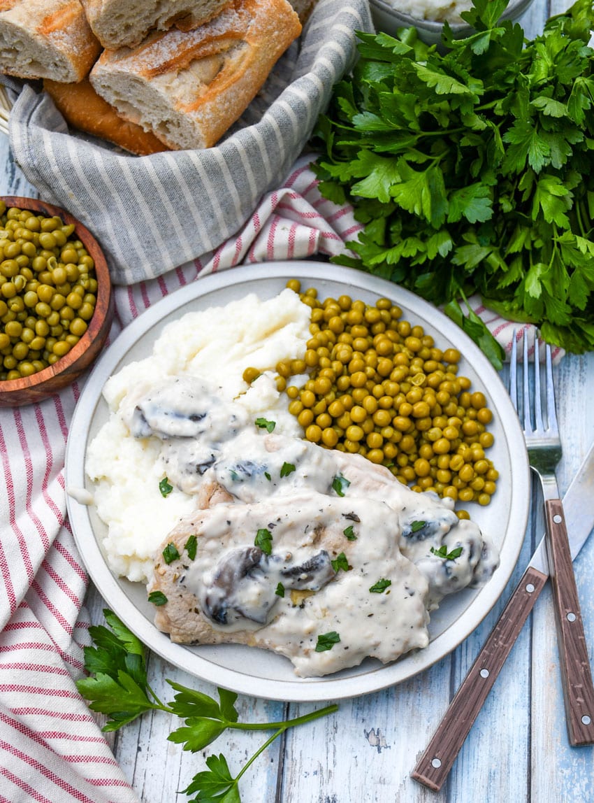 herbed pork chops in mushroom gravy over mashed potatoes with peas on the side on a white plate