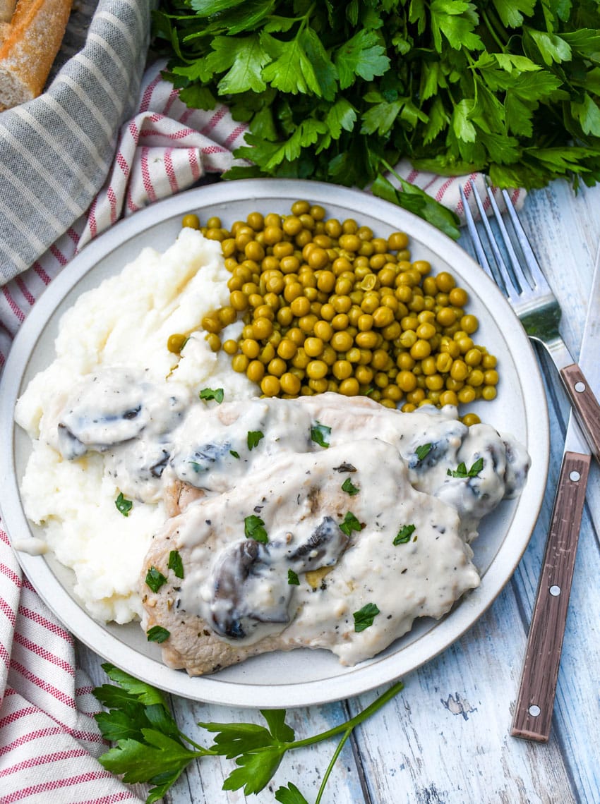 herbed pork chops in mushroom gravy over mashed potatoes with peas on the side on a white plate