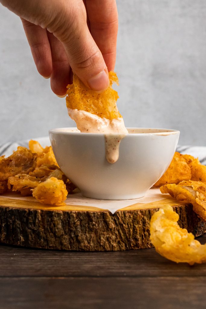 a hand dunking a bloomin onion bite into a white bowl of dipping sauce on a wooden cutting board