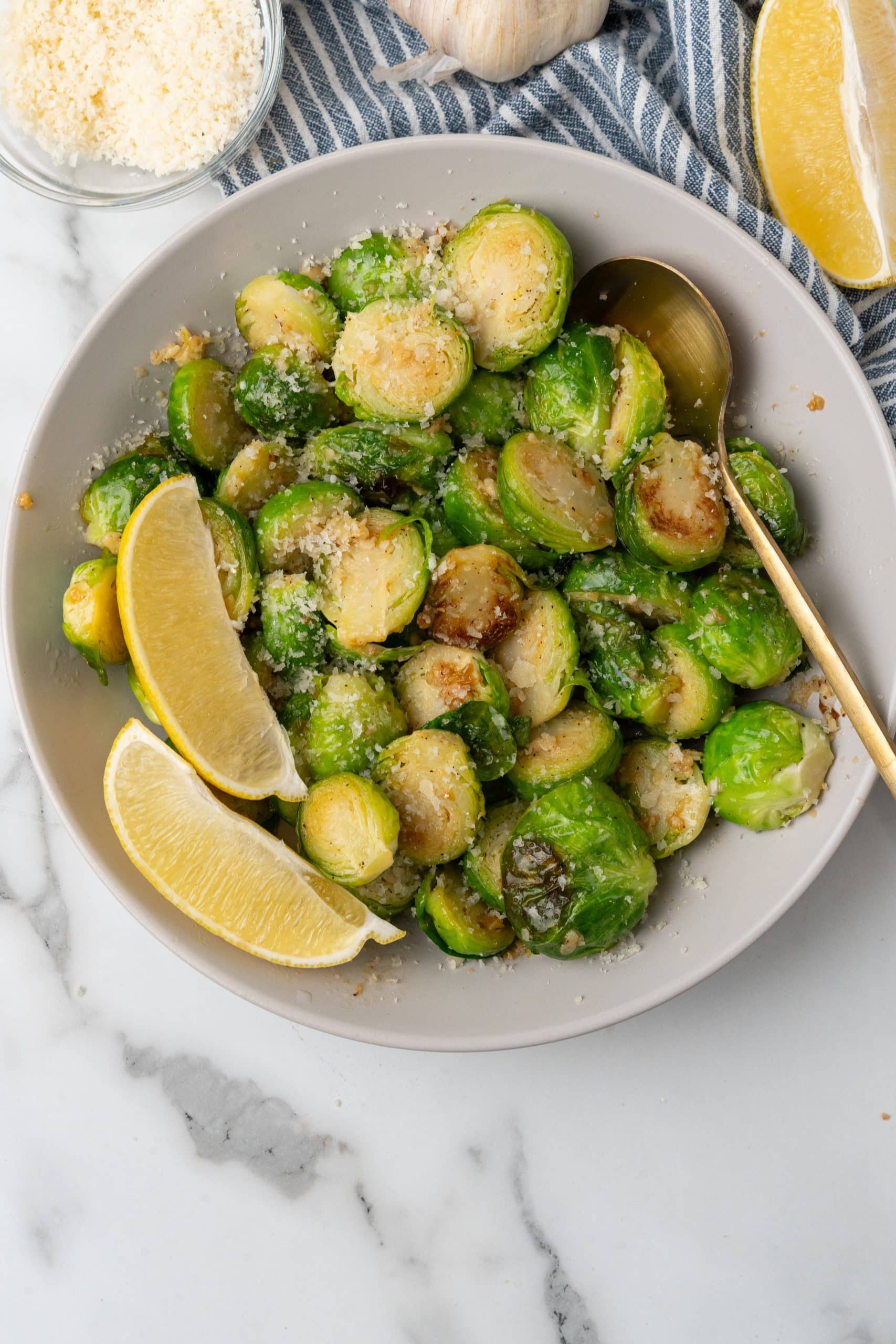 SAUTEED BRUSSELS SPROUTS WITH LEMON AND GARLIC in a white bowl sprinkled with parmesan cheese with lemon wedges on the side