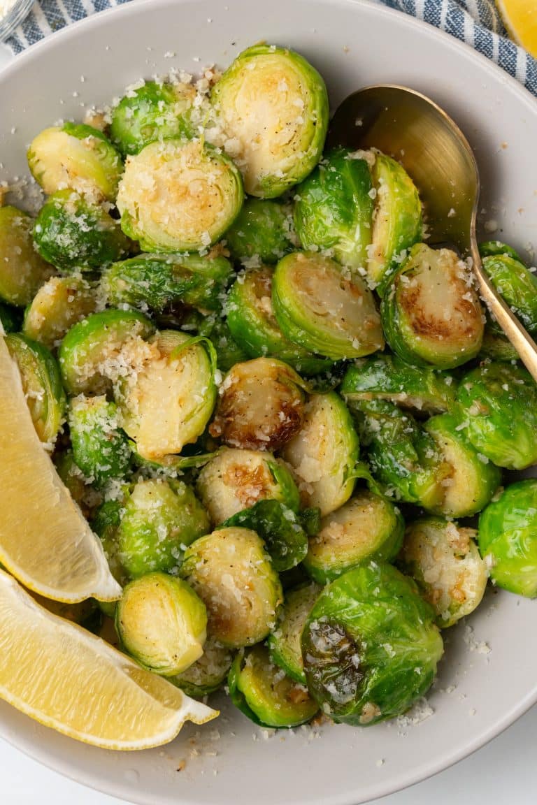 SAUTEED BRUSSELS SPROUTS WITH LEMON AND GARLIC in a white bowl sprinkled with parmesan cheese with lemon wedges on the side