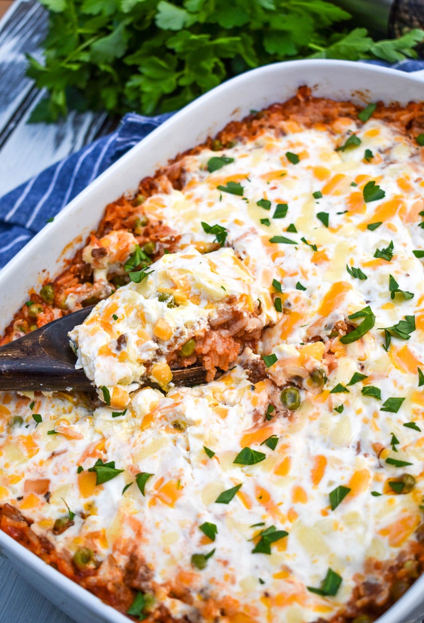 a wooden spoon scooping cheesy ground beef and rice casserole out of a white baking dish