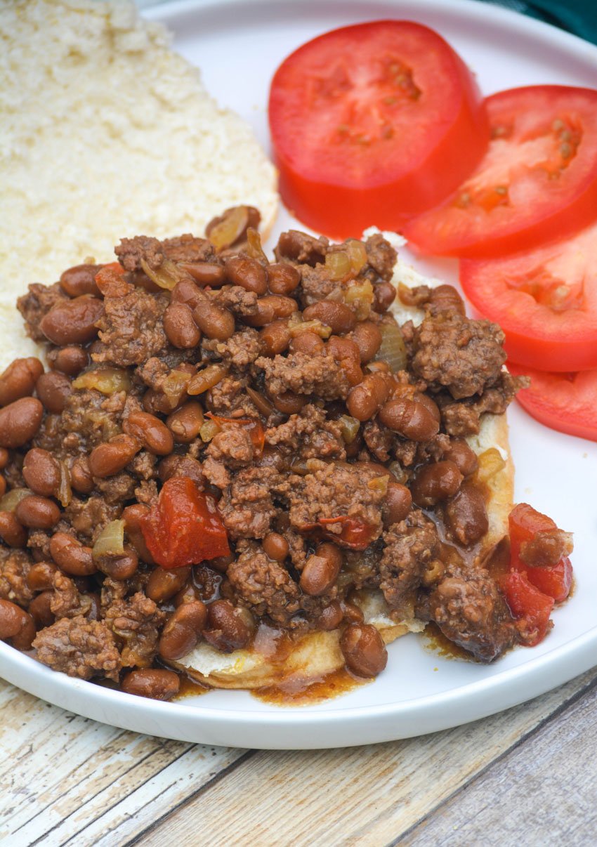 ground beef and baked bean cookout casserole served overtop a hamburger bun on a white plate with tomato slices