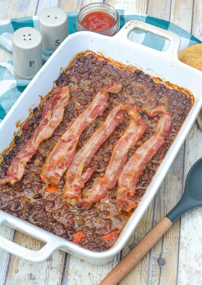 ground beef and baked bean casserole topped with sliced bacon in a white baking dish