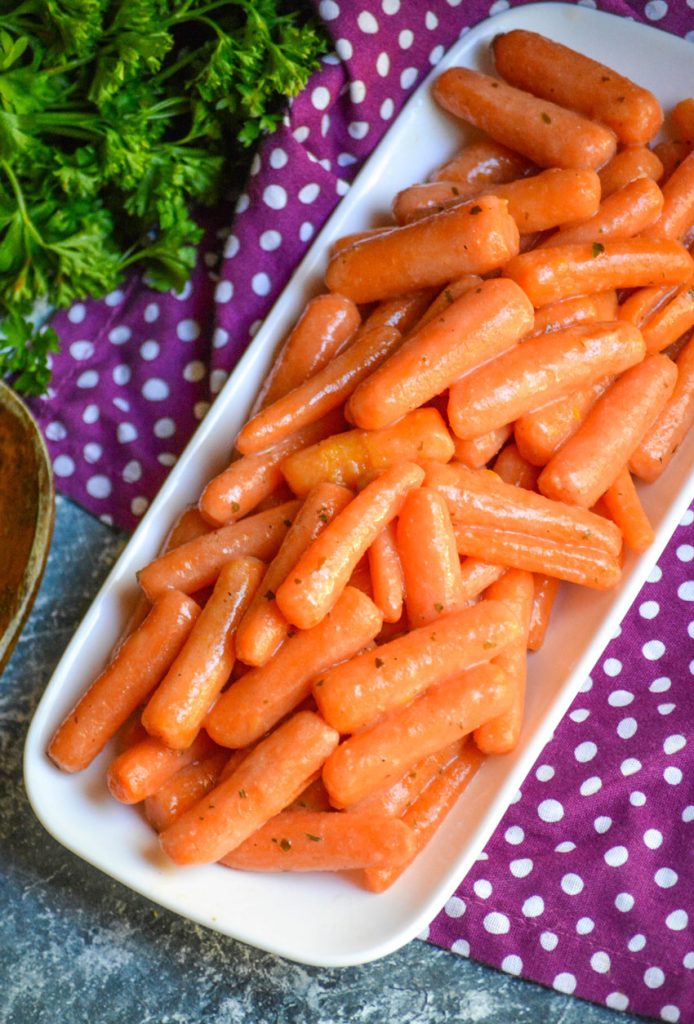 ranch glazed carrots piled on a skinny white serving dish with a wooden spoon and fresh herbs in the background