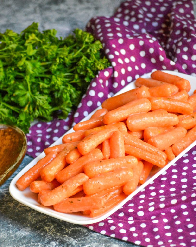 ranch glazed carrots piled on a skinny white serving dish with a wooden spoon and fresh herbs in the background