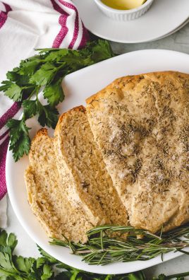 a loaf of crockpot rosemary & olive oil bread shown sliced on a white platter with fresh herbs on the side