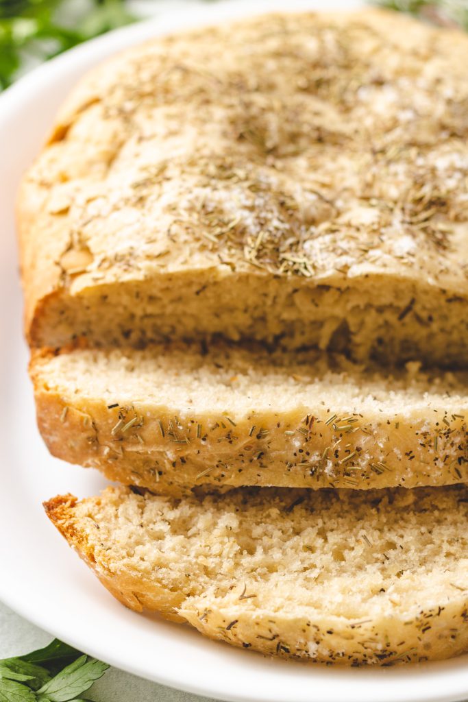 a loaf of crockpot rosemary & olive oil bread shown sliced on a white platter with fresh herbs on the side