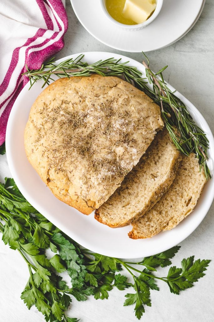 a loaf of crockpot rosemary & olive oil bread shown sliced on a white platter with fresh herbs on the side