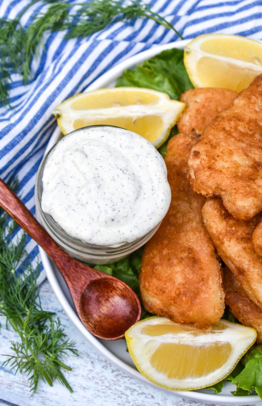 a small glass jar filled with homemade tartar sauce on a bed of lettuce next to fried fish on a white plate