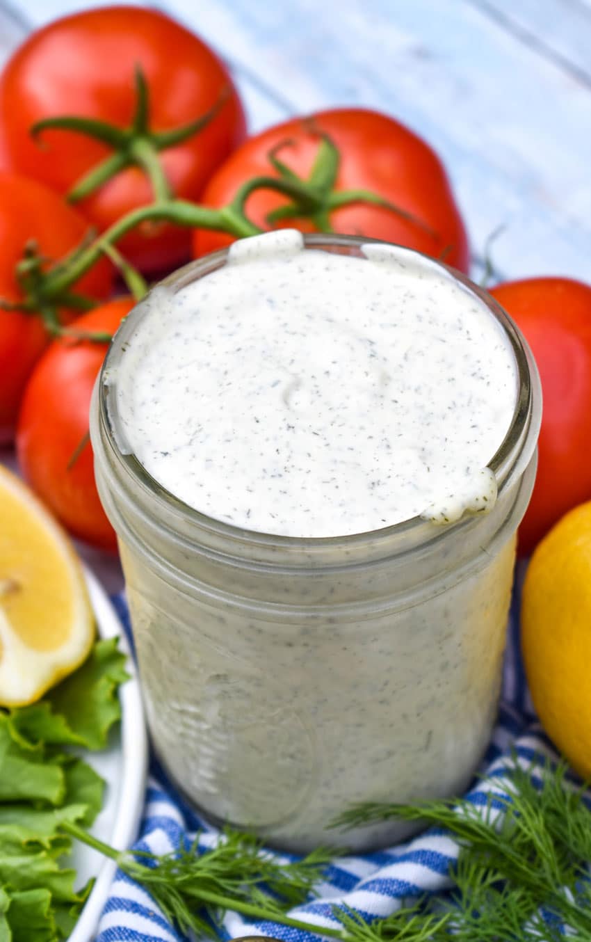 homemade tartar sauce in a glass jar surrounded by fresh herbs, tomatoes, and lemon wedges