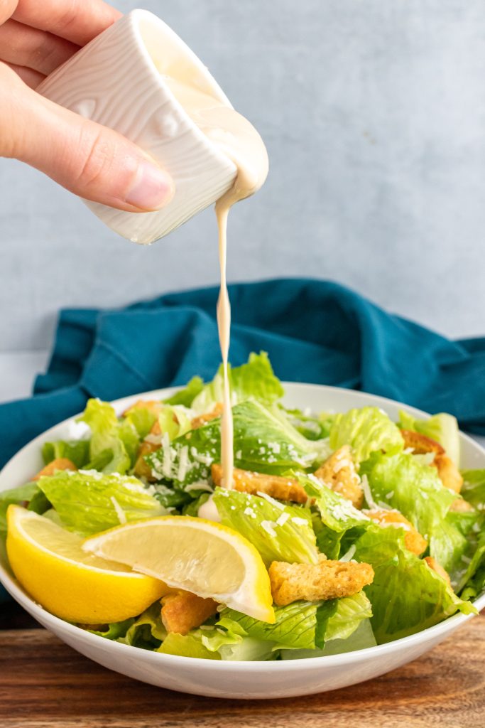 a hand shown pouring homemade caesar salad dressing over a bowl of salad