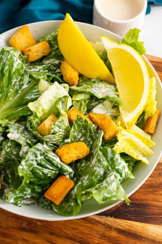 homemade caesar dressing tossed with lettuce in a white bowl shown topped with croutons, lemon wedges, and shredded cheese