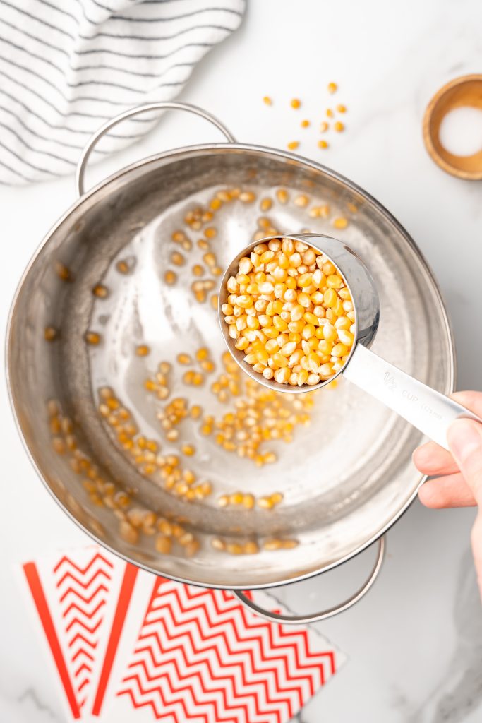 popcorn kernels being poured into a pot with hot oil and sugar