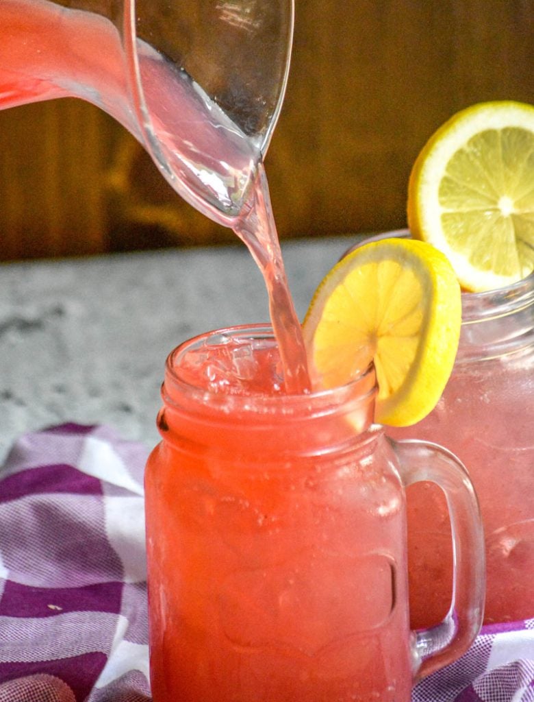 Blackberry Lemonade being poured from a glass pitcher into an ice filled glass jar garnished with a slice of lemon
