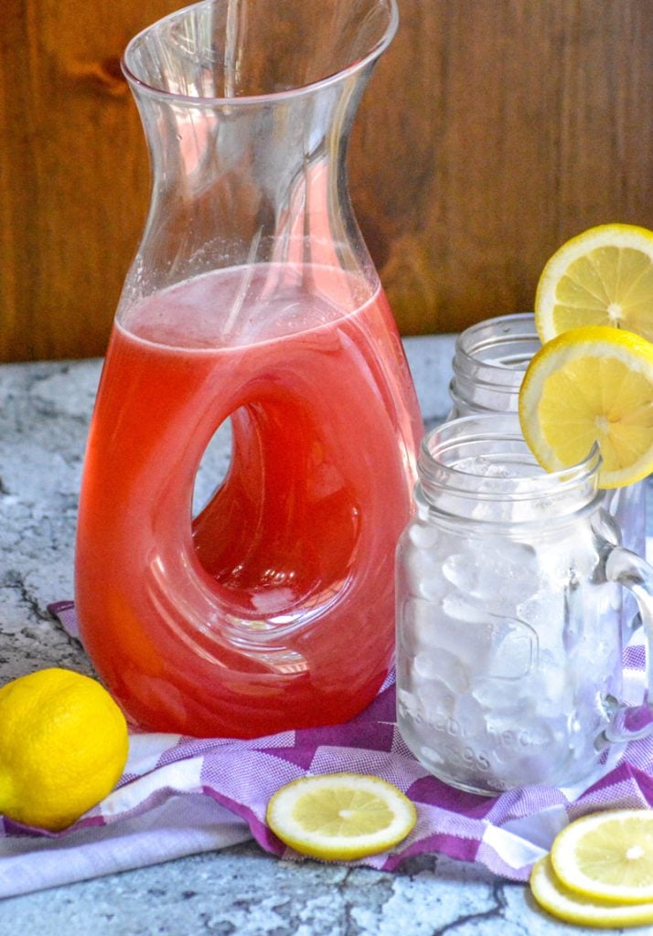 Blackberry Lemonade shown in a glass pitcher with sliced lemons and glasses filled with ice in the background