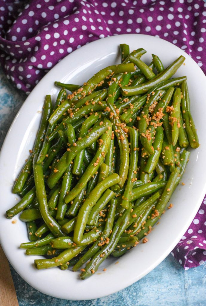 garlic teriyaki green beans in a white serving dish on a light blue background with a purple polka dotted cloth napkin in the background