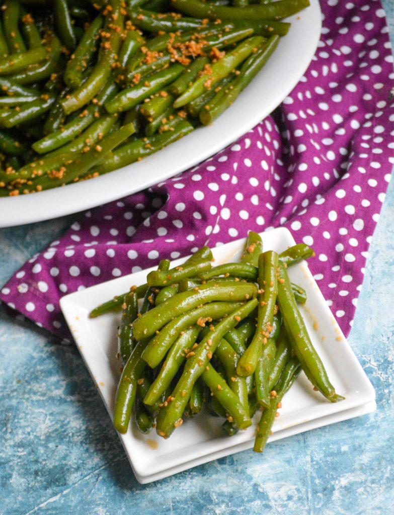 garlic teriyaki green beans piled high on square white plate with a purple & white polka dotted cloth napkin in the back ground