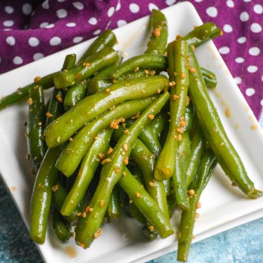 garlic teriyaki green beans piled high on square white plate with a purple & white polka dotted cloth napkin in the back ground