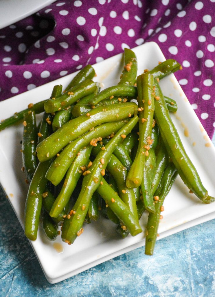 garlic teriyaki green beans piled high on square white plate with a purple & white polka dotted cloth napkin in the back ground