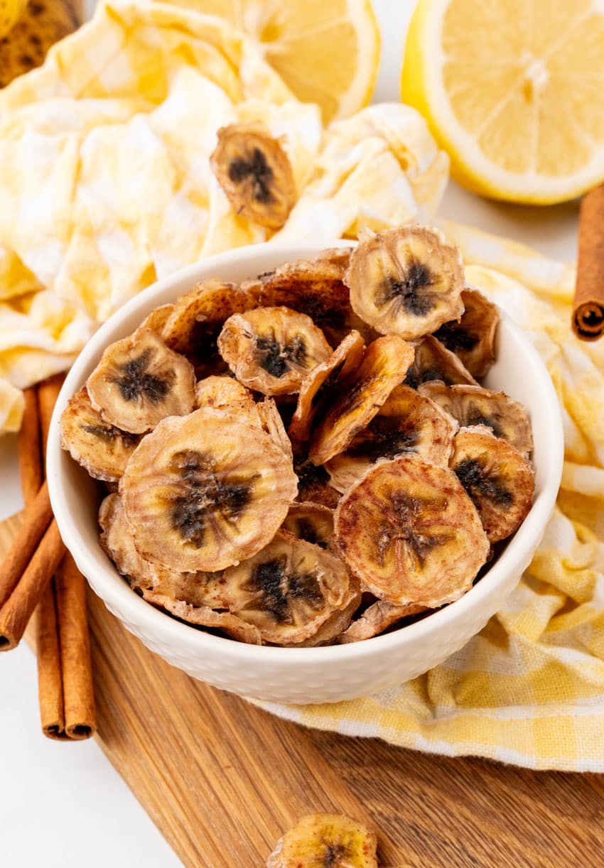 homemade banana chips in a small white bowl on a wooden cutting board